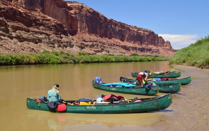 Five canoes rest on the shore of a river. A couple of people are getting things out of the canoes. The other side of the river is lined by a tall red canyon.